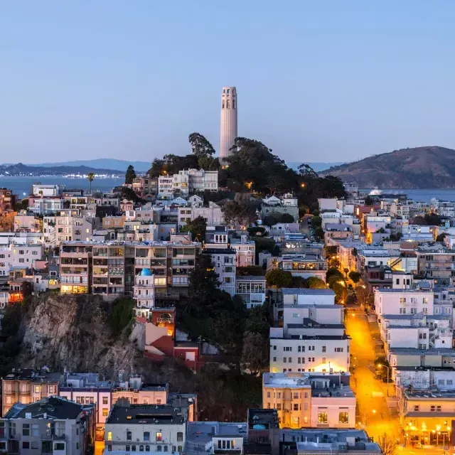 La Coit Tower di San Francisco al tramonto, con le strade illuminate davanti e la baia di San Francisco alle spalle.